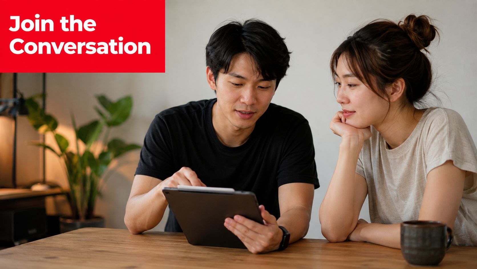 A young Asian couple sitting at a wooden table while looking together at a digital tablet screen.